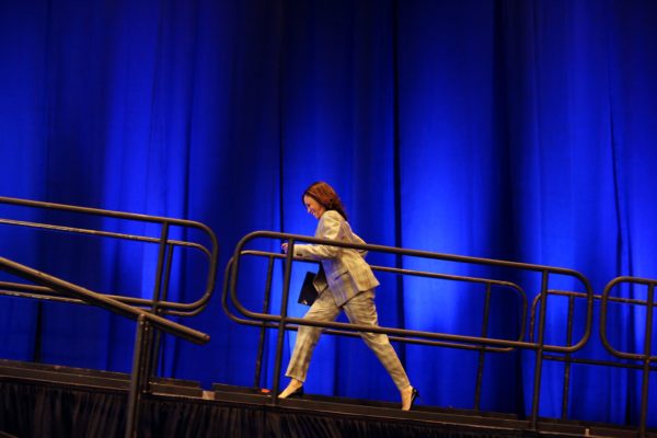 A picture of Kamala Harris walking up a ramp onto a stage. She is wearing a plaid pant suit and black high heels, smiling as she walks.