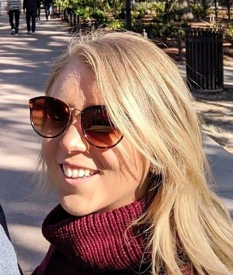 Katie has long blonde hair and is wearing sunglasses. She is on a street and wears a maroon turtleneck.