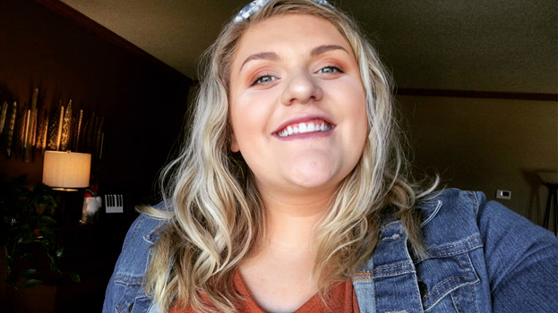 A picture of a woman with short, blonde hair smiling at the camera for a selfie. She is wearing a jean jacket with an orange shirt.