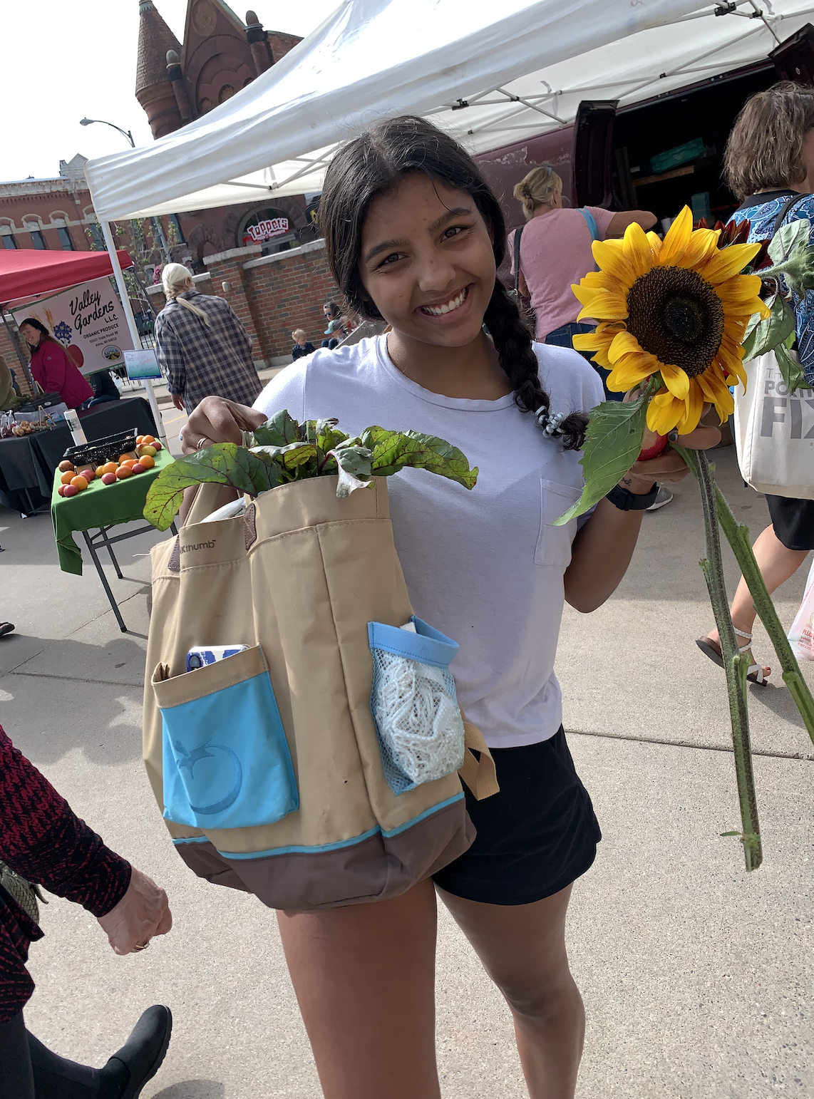 A picture of a woman in a white t-shirt smiling holding a bag of vegetables and a sunflower.