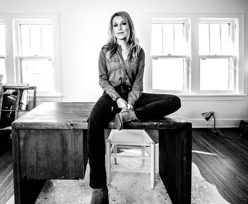 A black and white picture of a woman sitting on a wooden desk in a room. She is wearing a jean shirt with dark jeans.