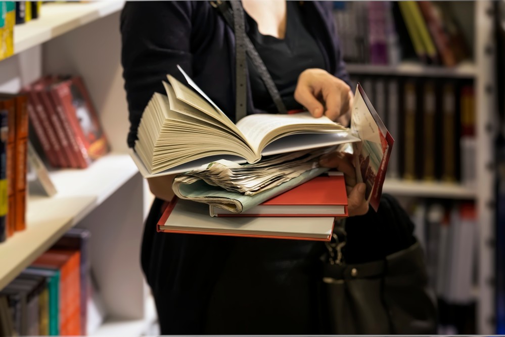 A picture of a woman holding a stack of books while reading the one that is on top.