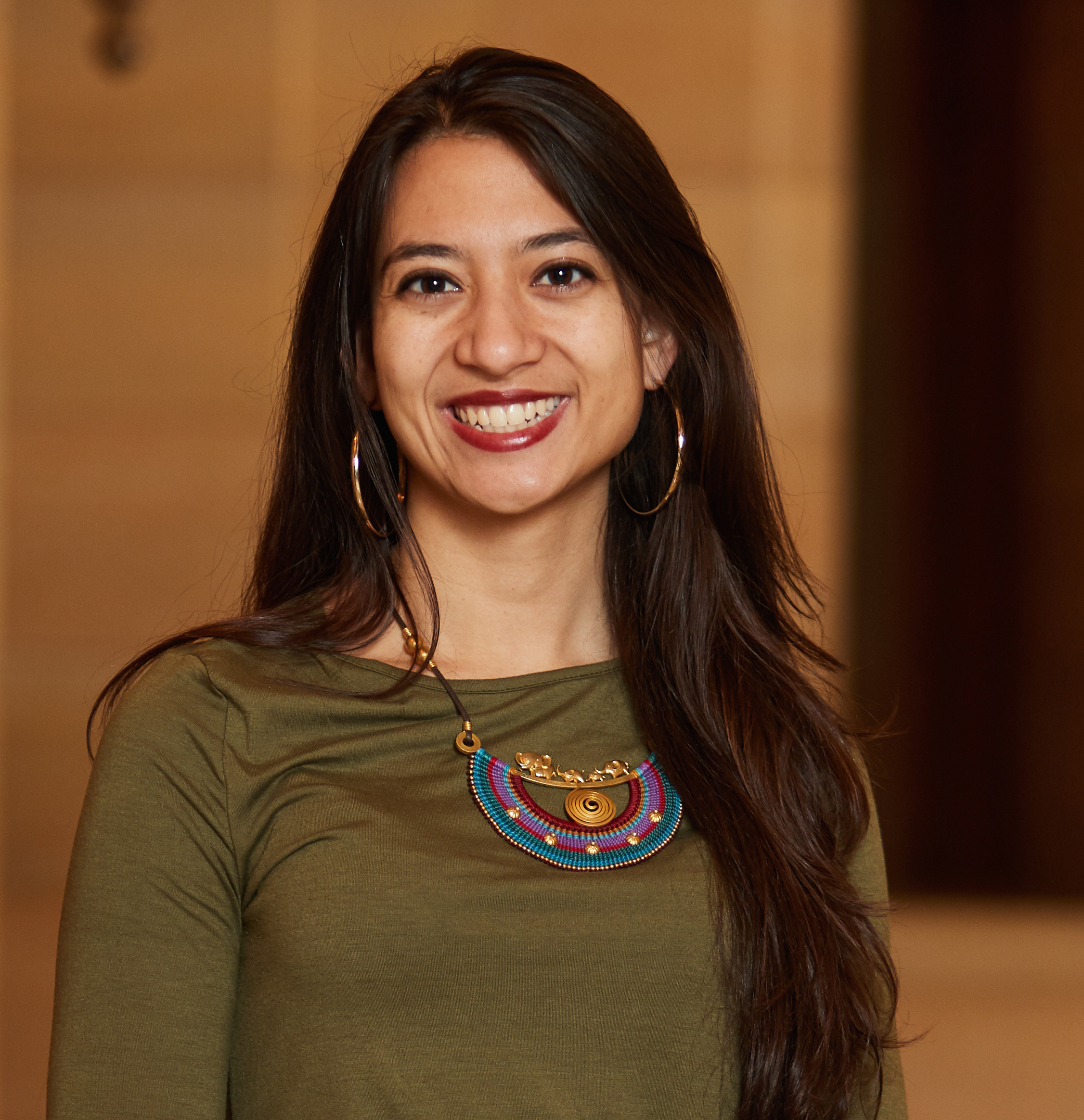 A picture of a woman with long dark hair smiling. She is wearing a green long sleeve, hoop earrings, and a decorated necklace.