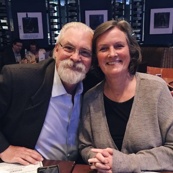 A picture of a woman and man together. The woman has short, dark hair and is wearing a black shirt and a brown cardigan. The man has glasses and facial hair, he has on a white button up and a black jacket.