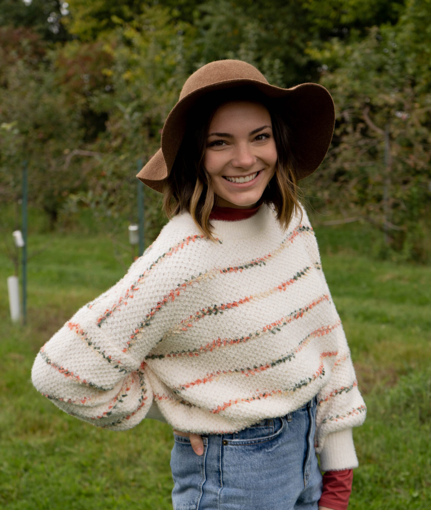 White woman with shoulder length brown hair. She is wearing a striped white sweater, blue jeans, and a lovely brown hair. She is standing in a green field.