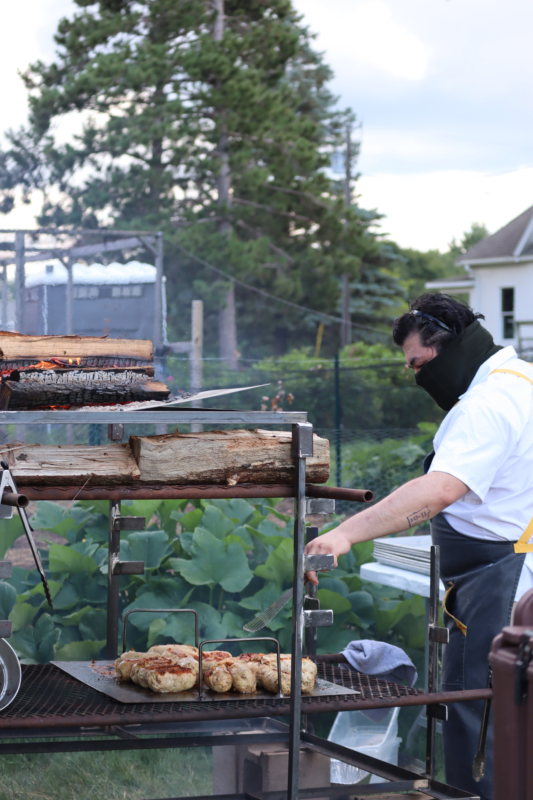 A picture of a man cooking meat over a grill. He is wearing a white shirt, a black apron, and a black face covering.