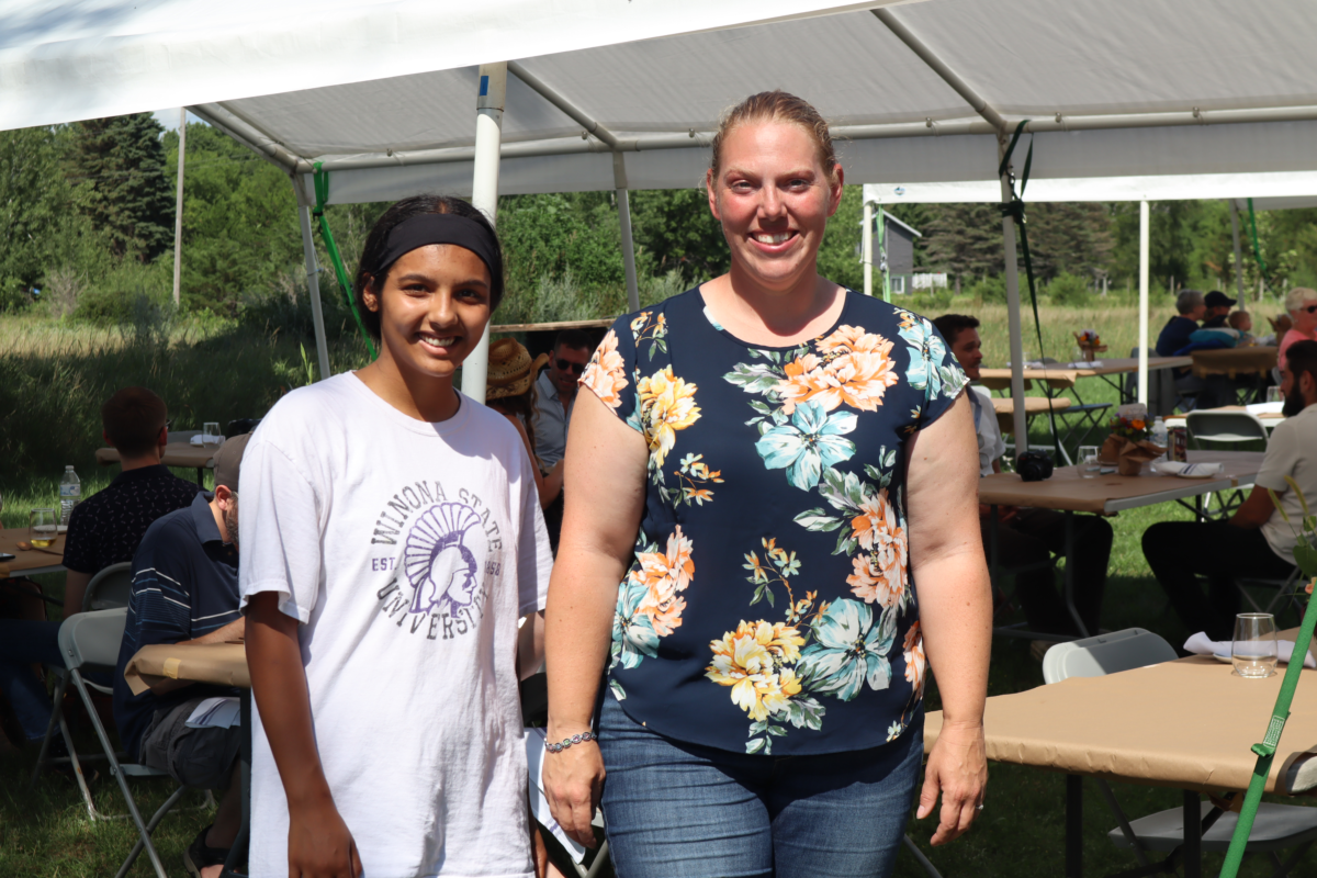 A picture of two women standing next to each other. One woman has dark hair pulled back into a ponytail and is wearing a white shirt. The other woman has blonde hair pulled back and she is wearing a floral shirt.