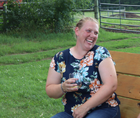 Carla is laughing while holding a warm drink on a park bench. She has light skin and blonde hair pulled up into a bun. She wears a blue floral shirt.