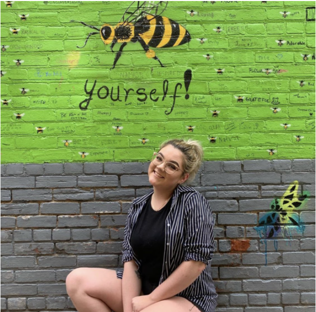 A picture of a woman with blonde hair wearing glasses, she's wearing a striped shirt and back t-shirt. Behind her is a painted mural on a brick wall.