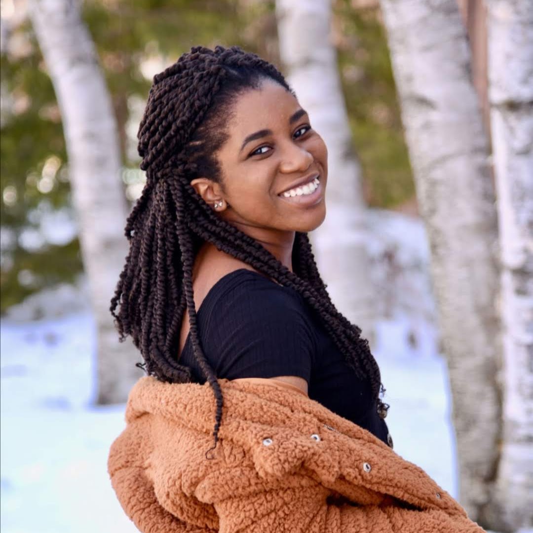 A picture of a black woman with long black hair looking back at the camera and smiling. She is wearing a black shirt with a brown fuzzy coat in front of a forest.
