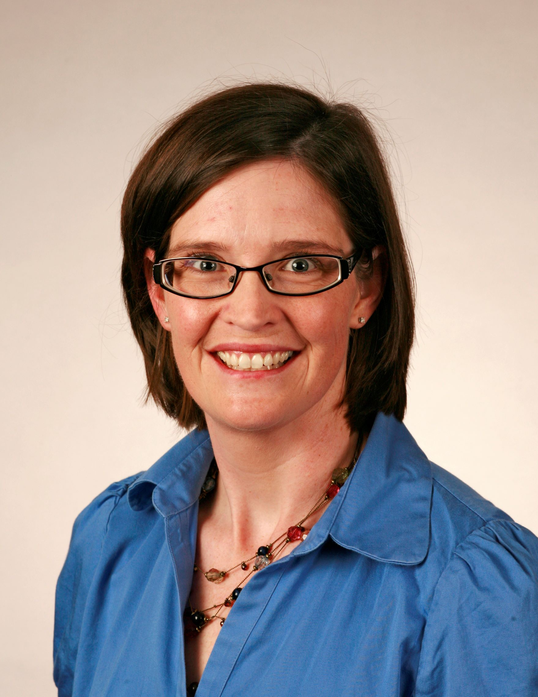 A picture of a woman with short, brunette hair with glasses smiling. She is wearing a blue blouse with a colored necklace.
