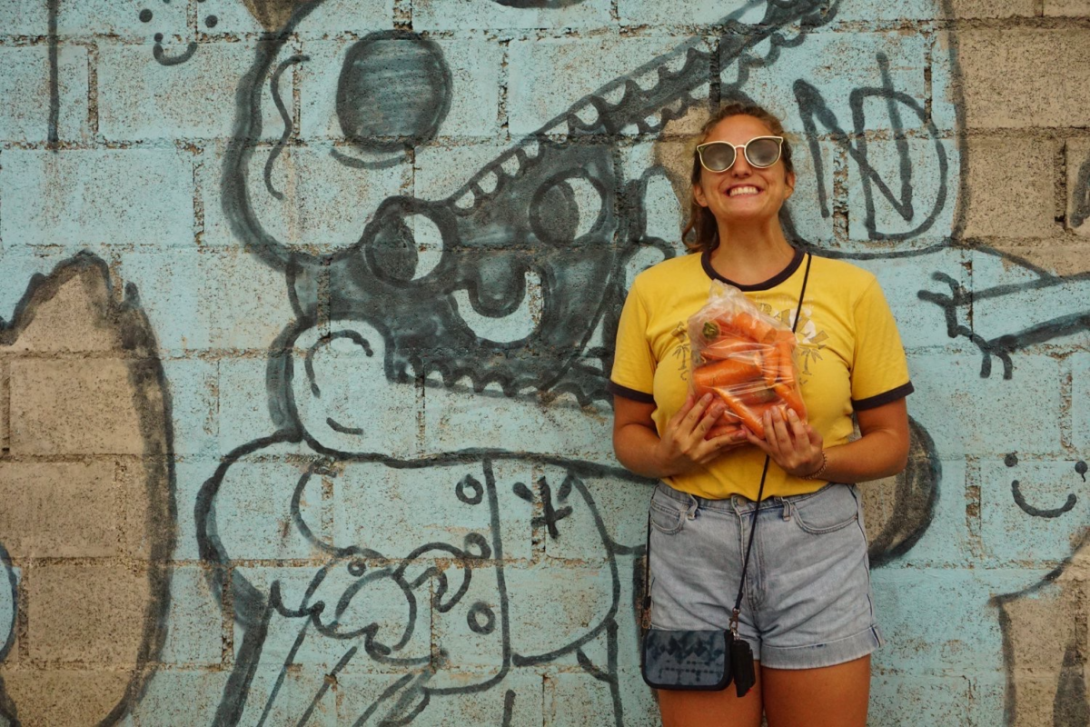 A picture of a woman with her hair pulled back and wearing sunglasses. She is wearing a yellow shirt and jean shorts, holding carrots in front of a painted mural.