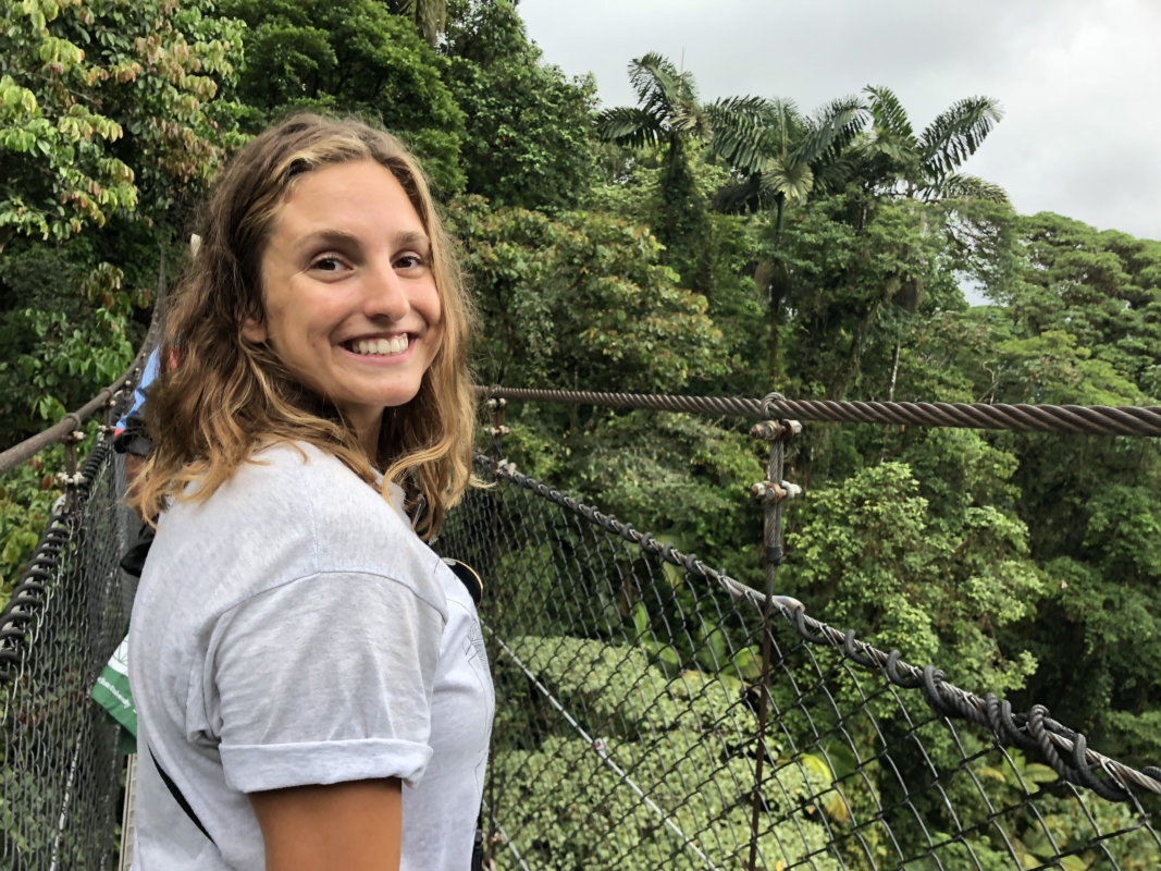 A picture of a woman with short, light hair on a bridge over a forest smiling.