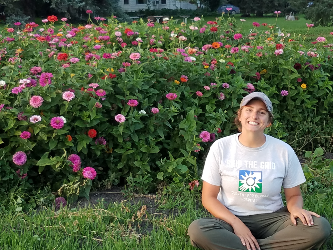 A picture of a woman with her hair pulled back and wearing a cap smiling. She is wearing a grey shirt and brown pants. She is sitting in front of flower bushes.