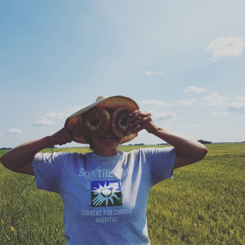 A picture of a woman wearing a grey t-shirt and sun hat holding two bagels up to her eyes in front of a field.