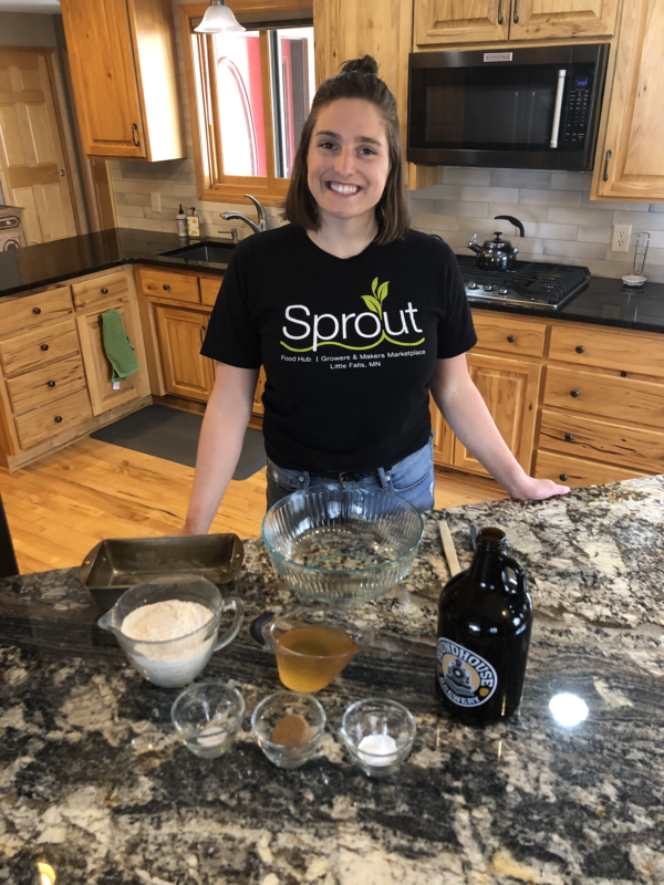 A picture of a woman with short, dark hair smiling wearing a black shirt that says "Sprout." In front of her are different ingredients as she stands in a kitchen.