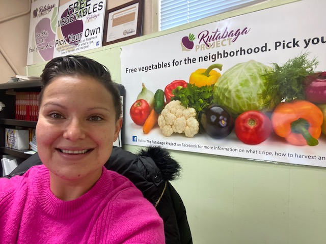 A picture of a woman with short brown hair wearing a pink sweater in front of a sign promoting vegetables.
