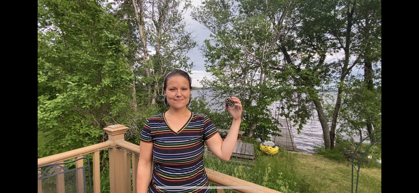 A picture of a woman with her hair pulled back in a pony tail wearing a striped v-neck. She is smiling and holding a pinecone while standing on a deck in front of a lake.