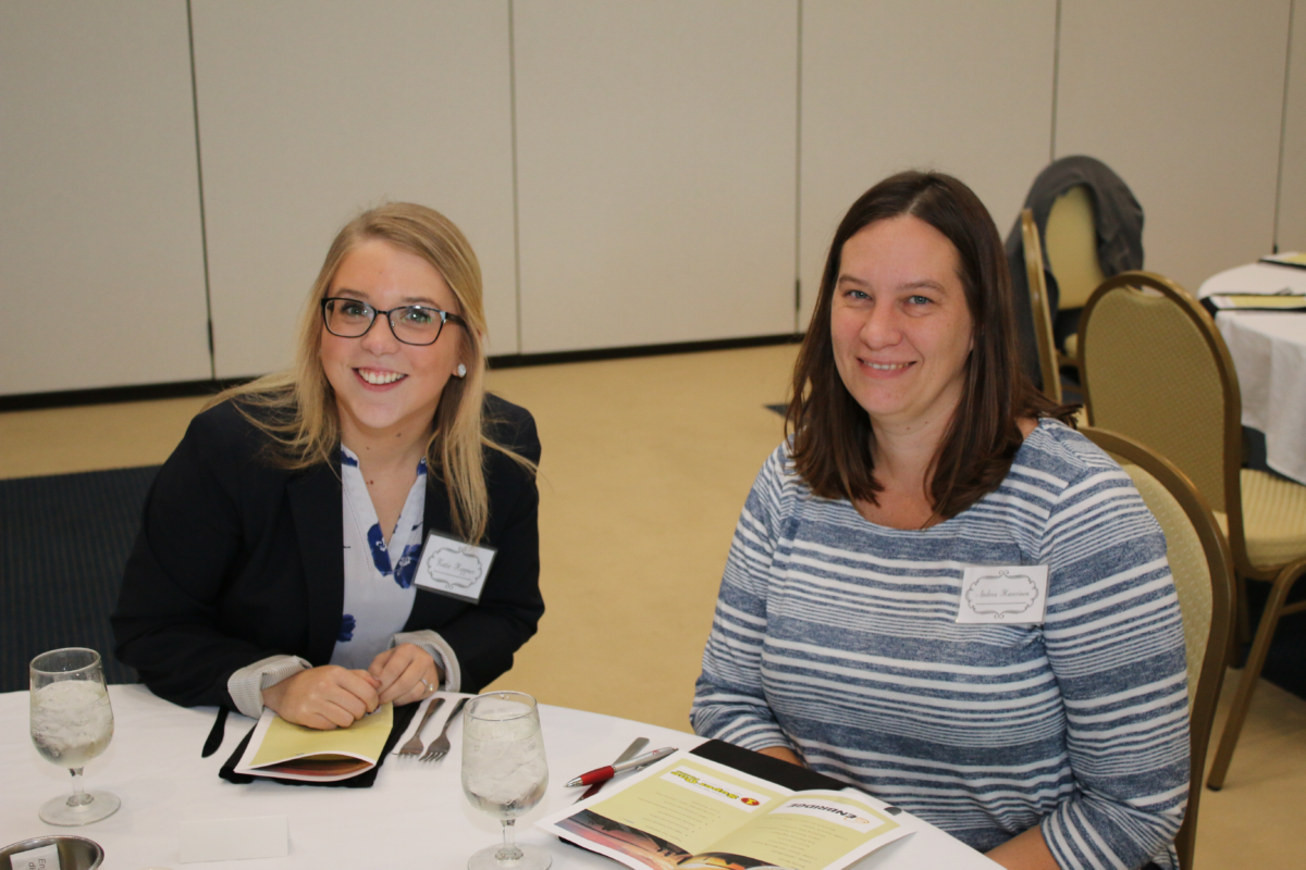 A picture of two women smiling and sitting at a table together. One woman has medium length blonde hair and glasses. She is wearing a black jacket and patterned white shirt. The other woman has short dark hair and is wearing a blue and white stripped shirt.