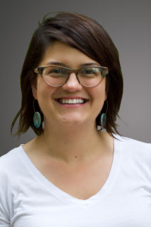 A picture of a woman with short, dark hair wearing glasses and smiling. She has a white v-neck on.