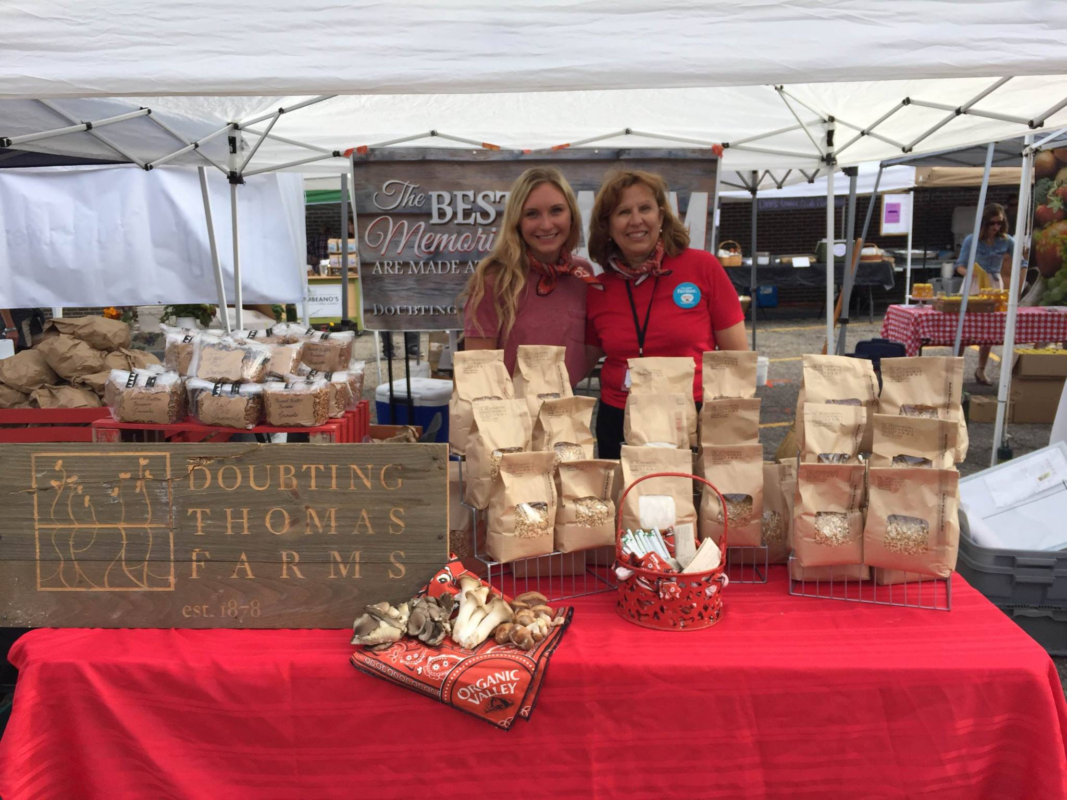 A picture of a young girl and woman at a farmer's market. The girl has long blonde hair and the woman has short red hair. Both are wearing red shirts to match the red table cloth. The sign reads "Doubting Thomas Farms, est. 1878"