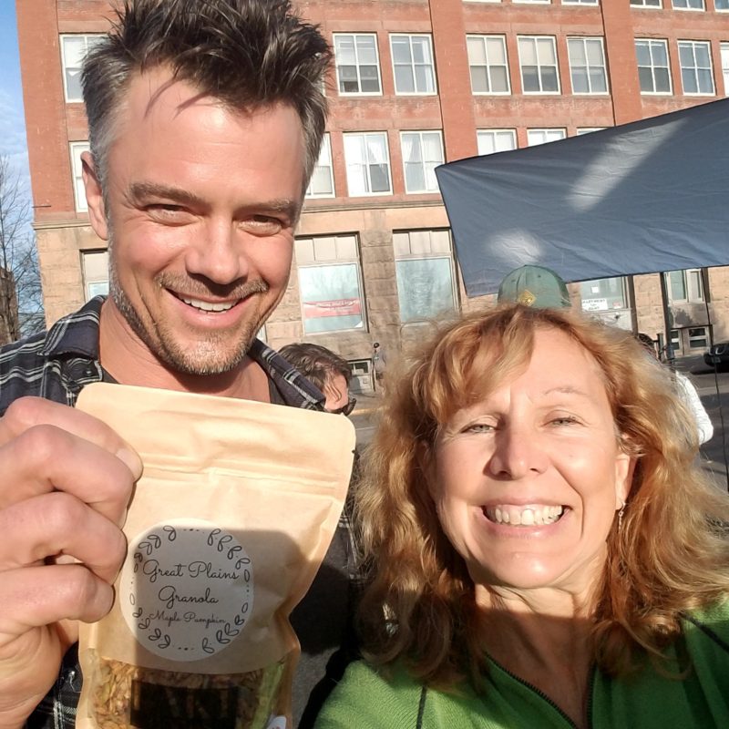 A picture of a man and a woman smiling. The woman has short, curly red hair and is wearing a green shirt. The guy has short, dark hair with some grey and is wearing a plaid shirt. He is holding some granola from a farmer's market.