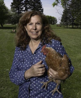 Noreen has medium length curly brown hair and is smiling while holding a chicken. She has light skin and is wearing a blue polka dot button up.