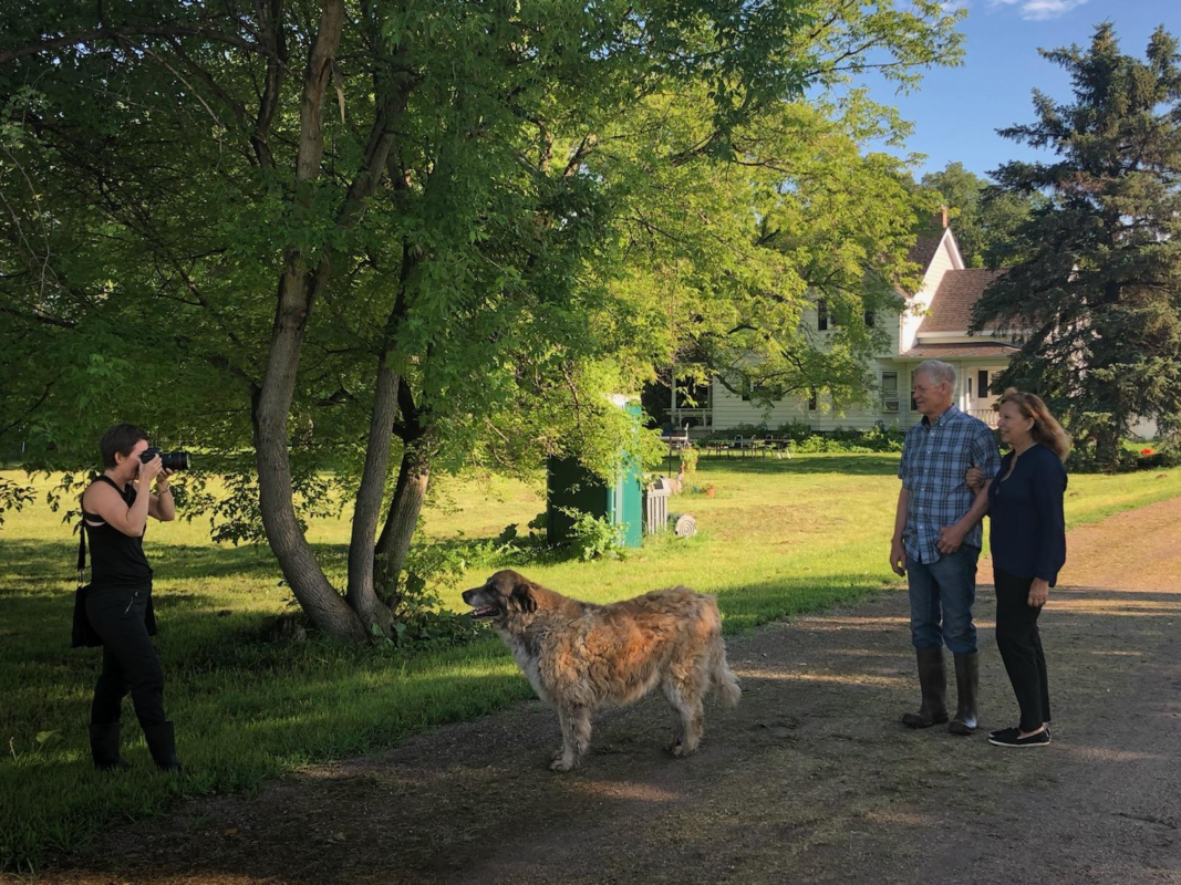 A picture of a photographer taking a picture of a man and woman on a farm. A dog is shown between the couple and the photographer.