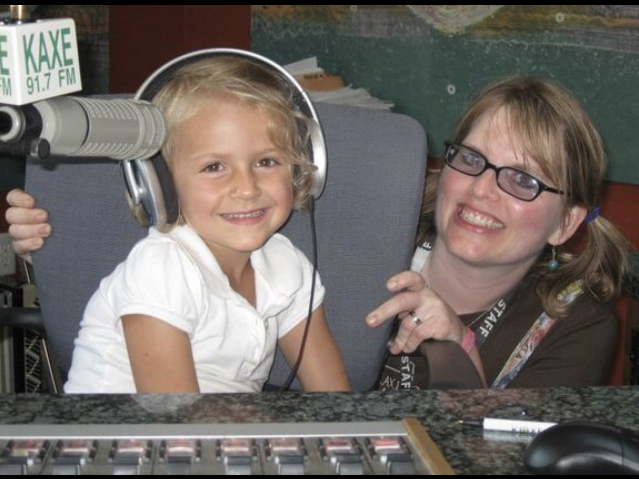 A picture of a little girl with an older woman. The little girl has blonde hair and is wearing a headset and a white polo. The woman has blonde hair in pigtails and glasses, she is wearing a brown shirt.