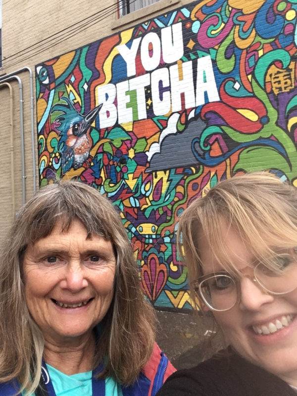 A picture of two women in front of a mural that says "You Betcha." The woman on the left has brown hair with bangs and is smiling. The woman on the left has blonde hair and glasses and is also smiling.