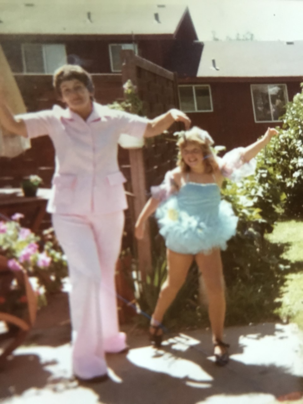 A picture of a girl with blonde hair in a blue dance costume with her grandma who is wearing a pink jumpsuit in front of their house.