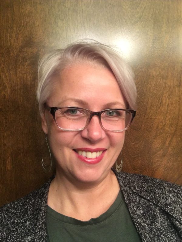 White woman with very light blonde short hair. She is smiling, wearing a green and grey blouse with black horn-rimmed glasses.