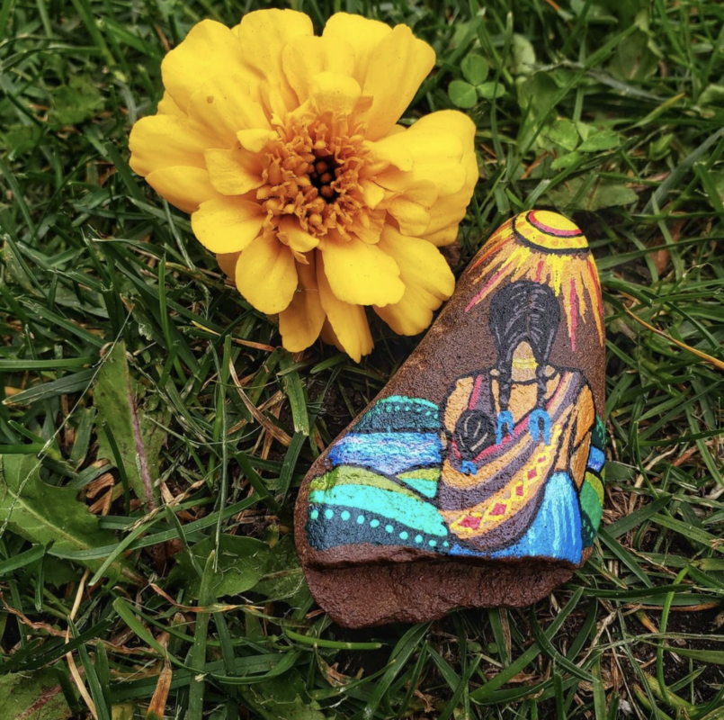 A picture of a yellow flower next to a painted rock on the grass. The rock shows a Native America looking out over a field with a child in her sling.