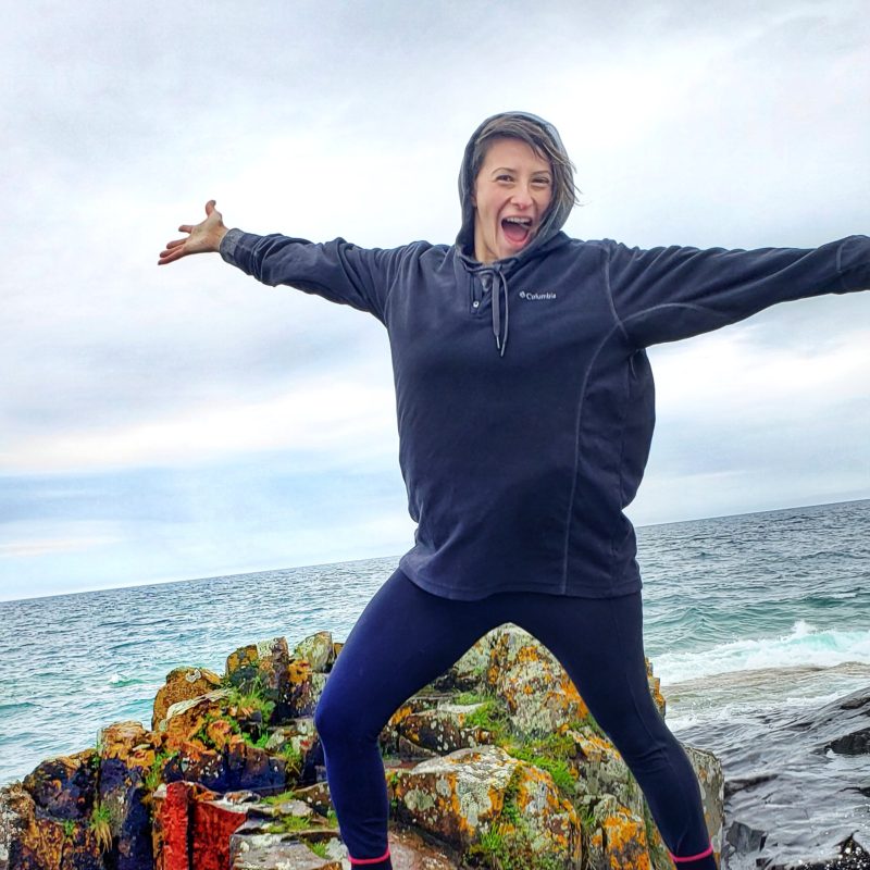 A picture of a woman next to one of the Great Lakes with her arms stretched out. She is standing on some rocks and has short brown hair, wearing a dark colored covering and sweatshirt.