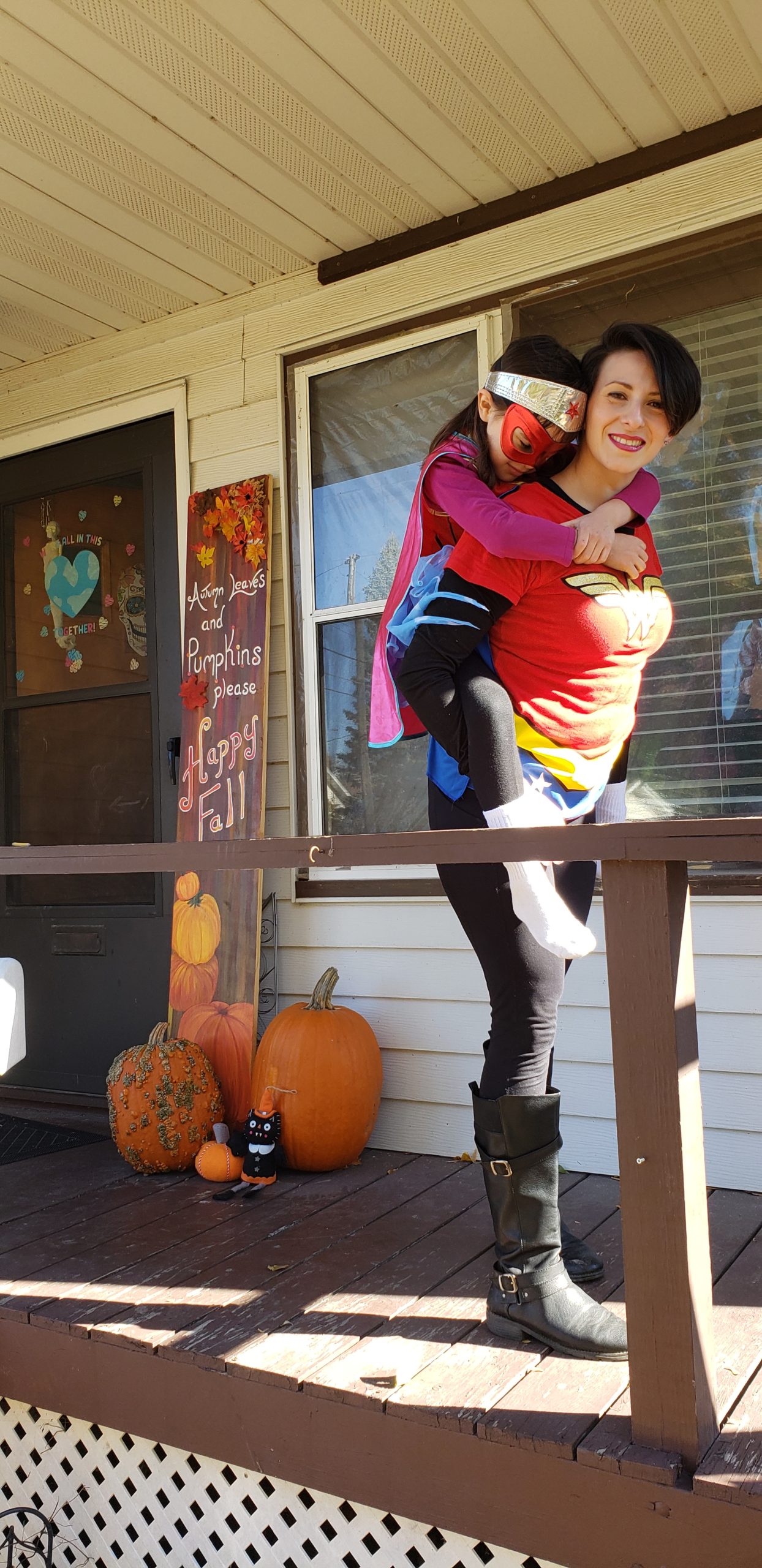A picture of a woman with her child on her back in fall time. In the back are pumpkins and fall decorations.