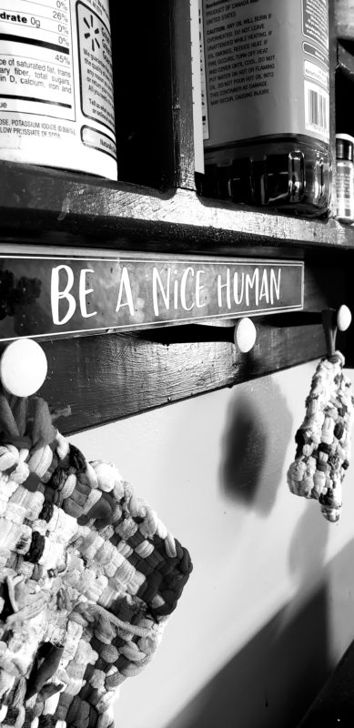 A black and white picture of a sign saying "Be A Nice Human" on a hook rack.