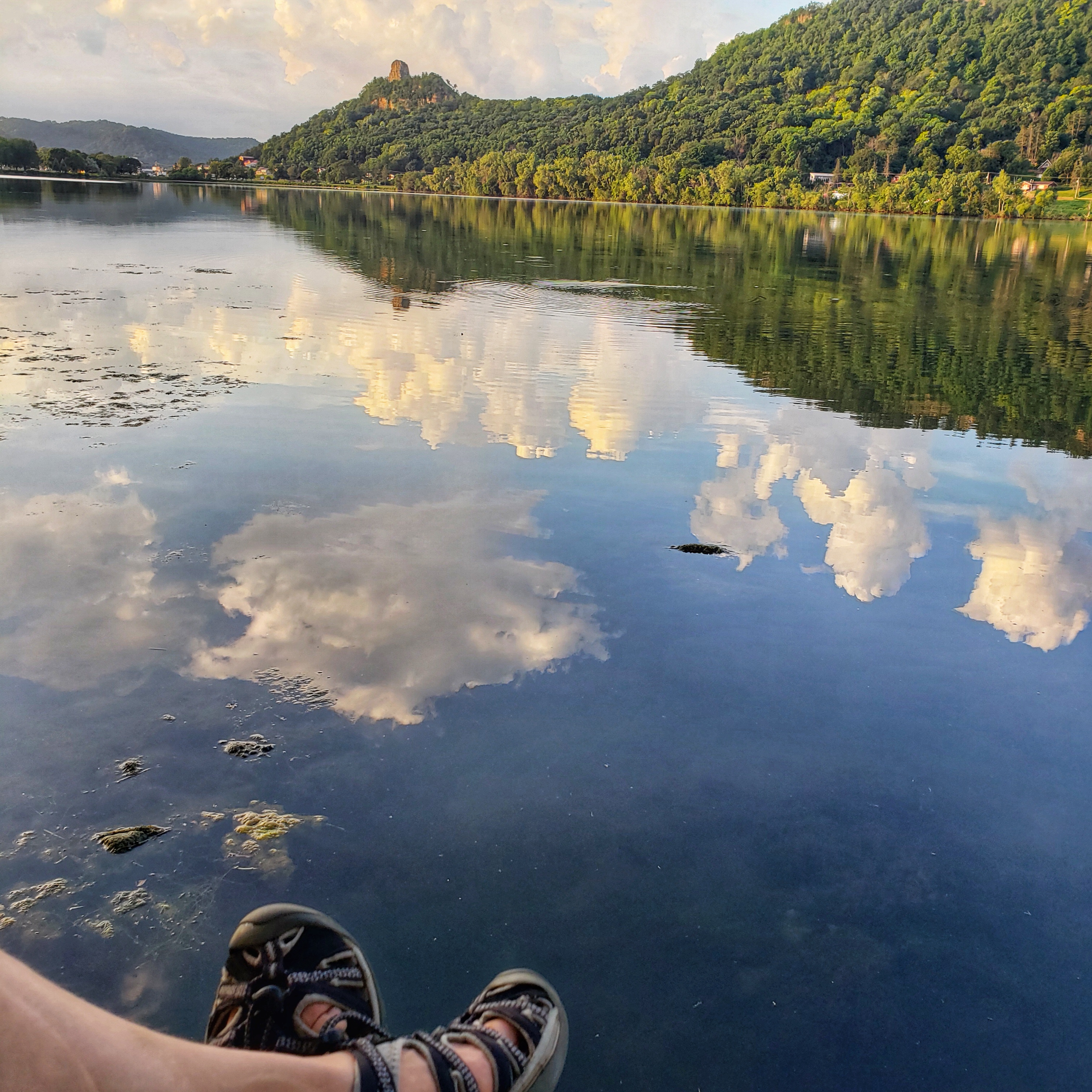 A picture from Winona, Minnesota that displays Sugar Loaf Bluff and Lake Winona. A pair of feet can be seen resting over the water.