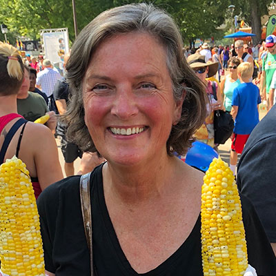 A picture of a woman with short grey hair wearing a black shirt smiling. She is at a fair holding two sweetcorns.
