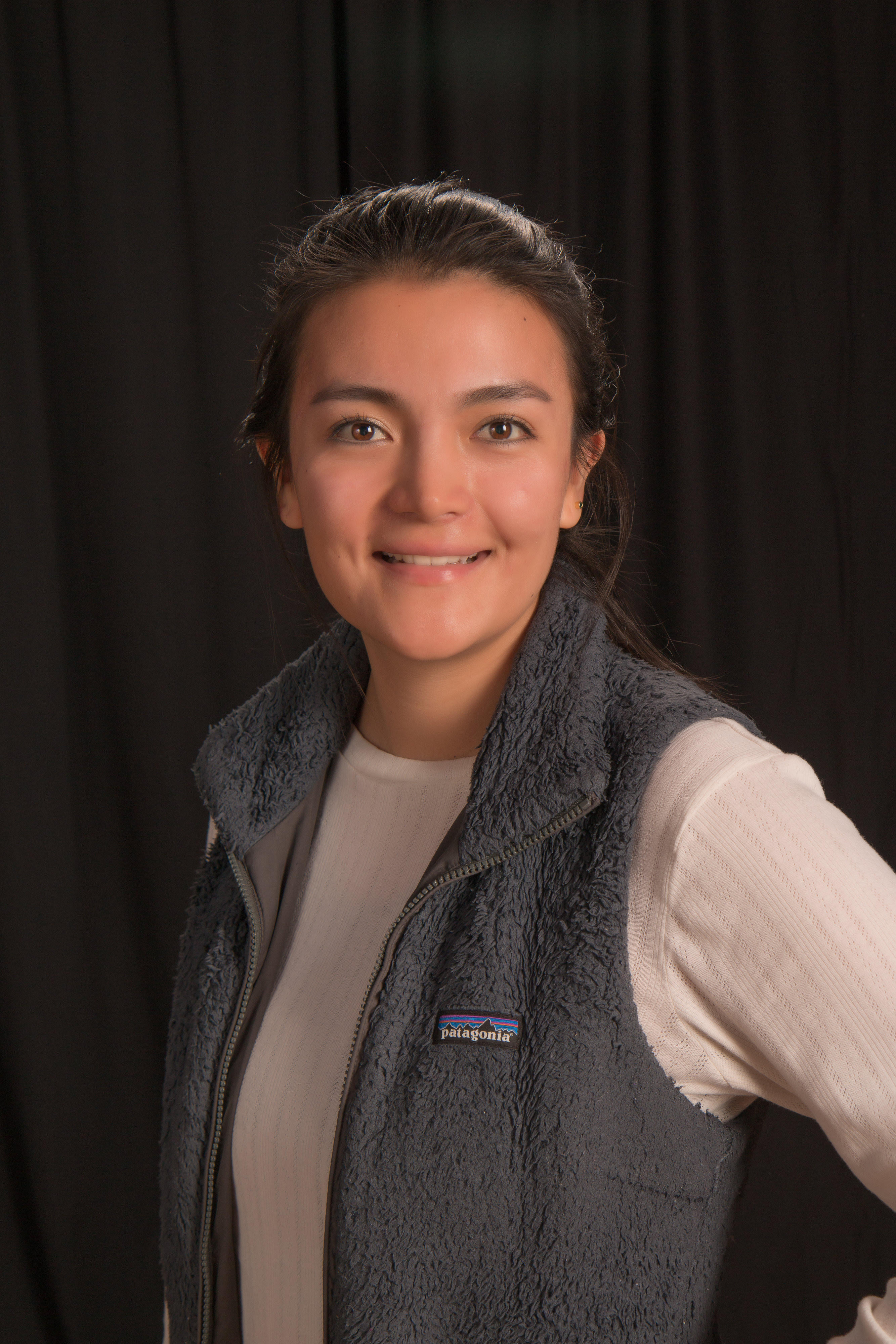 A picture of a woman with dark hair pulled back wearing a grey vest and white long sleeve smiling.