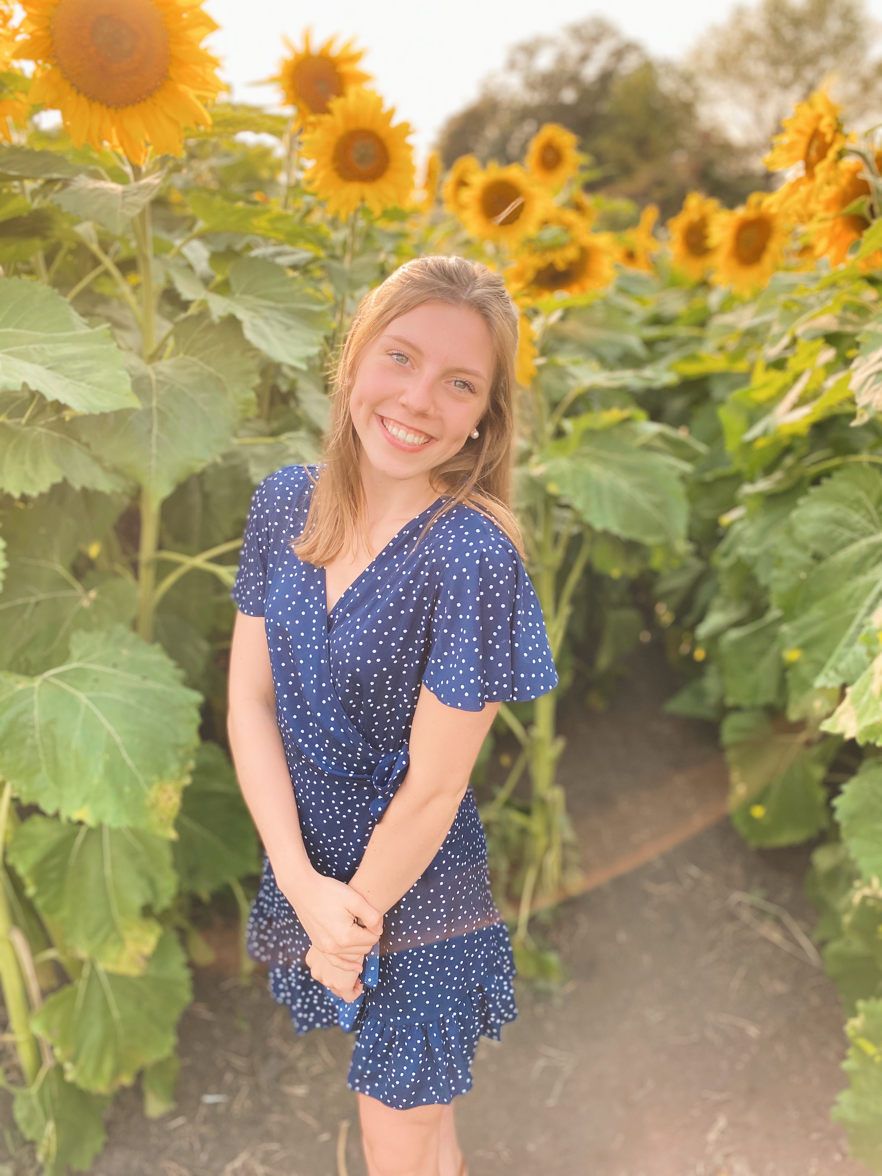 A picture of a smiling girl with brown hair in front of a sunflower field.