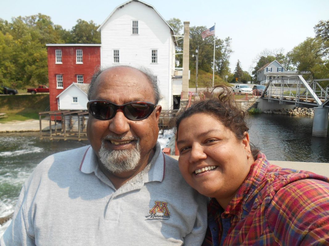 A picture of a man and woman smiling near a river. In the background are an old river factory, house, and bridge.