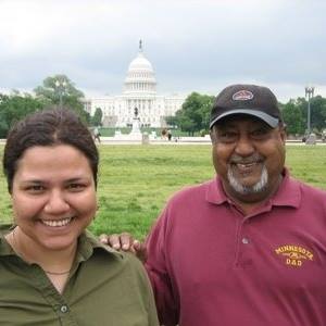 A picture of a smiling man and woman in front of the United States capital. The woman has dark hair and is wearing a green shirt. The man is wearing a hat and has facial hair, he is wearing a maroon polo.