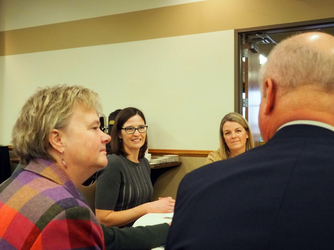 A picture of four people at a table meeting. Two women are shown smiling, one with short dark hair and glasses; the other has longer blonde hair. The woman closet to the camera has short dark hair and displays an engaged posture.