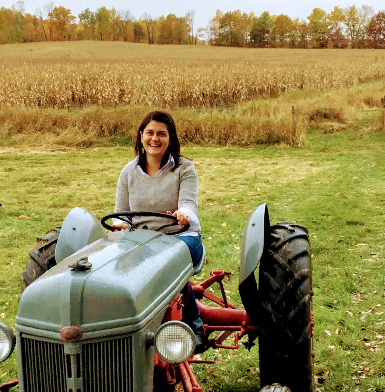 A picture of a woman with short dark hair smiling and sitting on a tractor in a field.