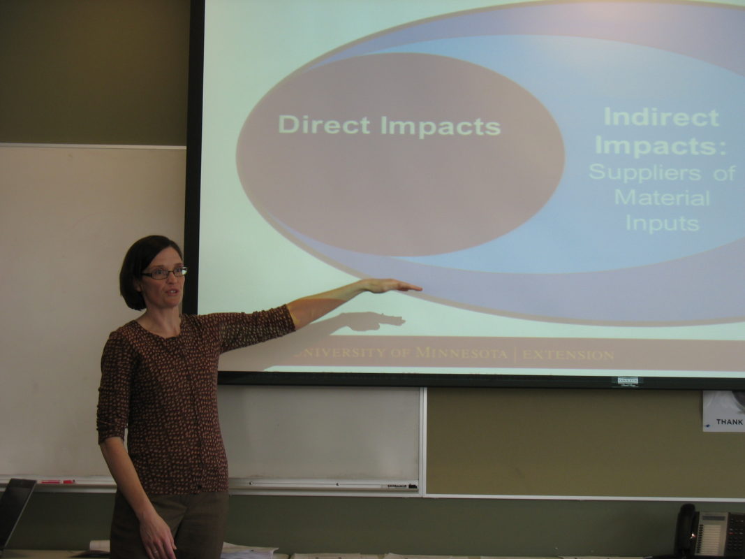 A picture of a woman with short brown hair and glasses wearing a maroon patterned shirt giving a powerpoint presentation about direct and indirect impacts.
