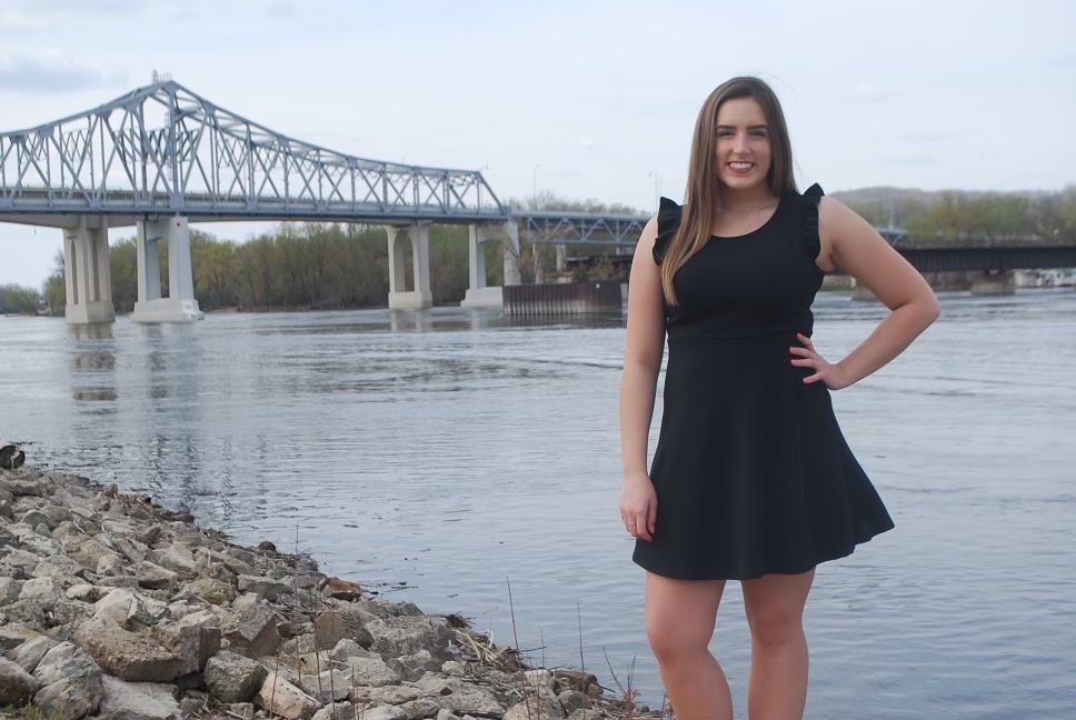 A picture of a woman with long brunette hair wearing a black dress smiling in front of the Mississippi River and a bridge.