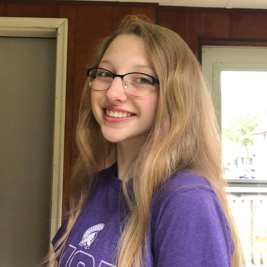 A blonde woman with glasses wearing a purple shirt smiling.