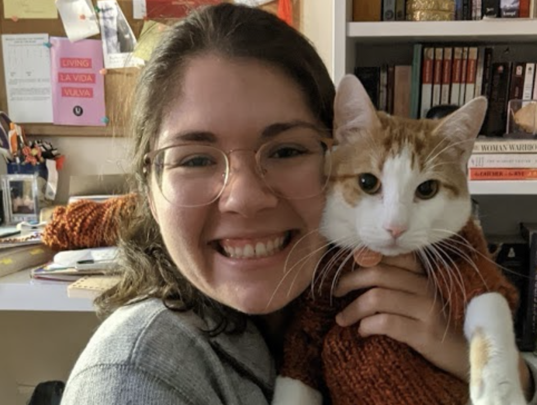 A picture of a woman with short dark hair and glasses smiling and holding a car. She is wearing a grey long sleeve and the cat is wearing a orange sweater.