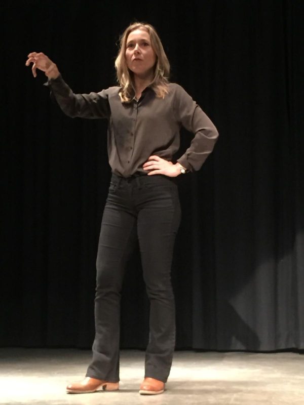 A picture of a blonde woman presenting on stage. She is wearing a brown long sleeve button-up and black pants with brown boots.