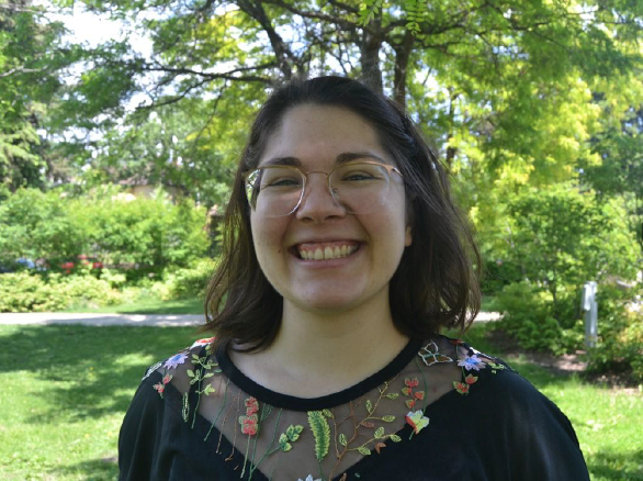 A picture of a woman with short dark hair and glasses smiling. She is wear a dark shirt with floral prints and is standing in front a green space.